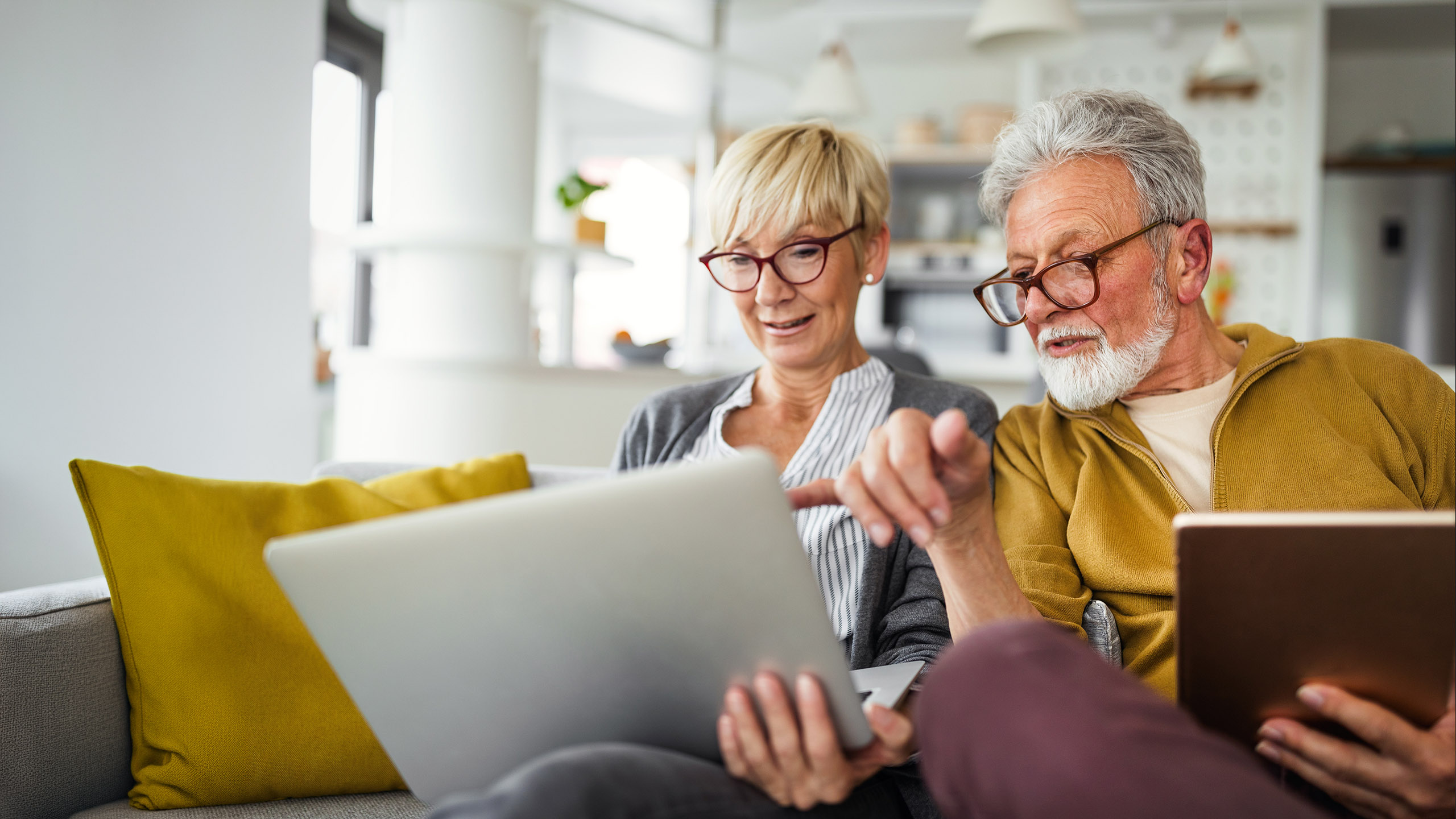An older couple sits on a grey sofa, looking at a silver laptop. The man, with a white beard and glasses, points at the screen while the woman, with blonde hair and glasses, holds the laptop. A brown tablet is also visible in the man's lap.