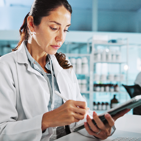 A woman in a white lab coat uses a stylus on a digital tablet in a lab.