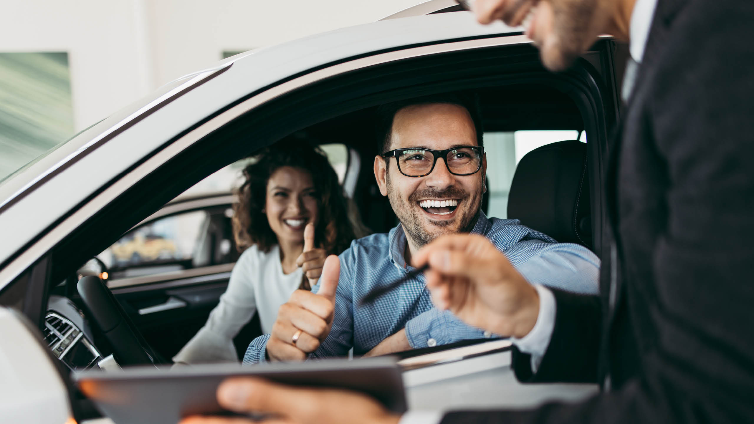 A smiling man and woman in a new car talking to car salesman, who is holding a tablet. The man holds a pen, ready to sign.