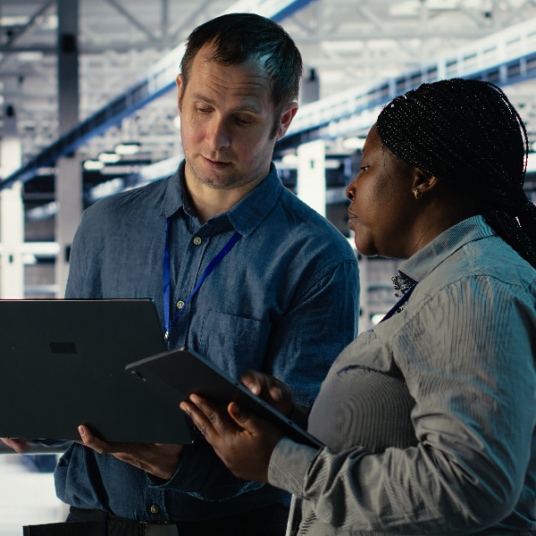 A man and a woman, both wearing lanyards, look at a laptop and tablet respectively in office.