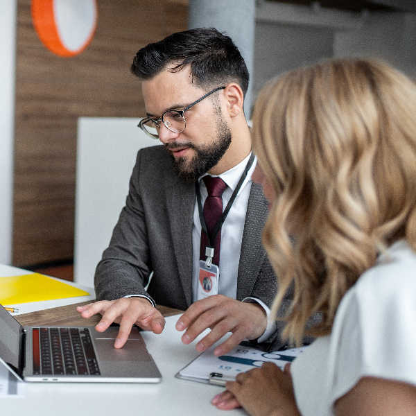 A man in a suit and glasses works on a laptop next to a woman looking at a clipboard at a desk.