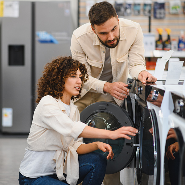 A man and a woman in an appliance store looking at a front-loading washing machine or dryer. The woman crouches, touching the open door, while the man stands, looking at the controls.