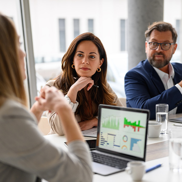 Three professionals in a meeting, two listening intently while a laptop displays data visualizations on a white table.