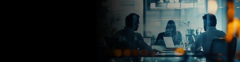 Three people working on laptops in a dimly lit meeting room, seen through a reflective glass partition.