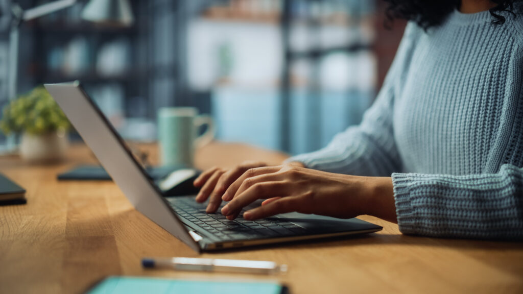Close-up of a person's hands typing on a laptop keyboard at a wooden desk.