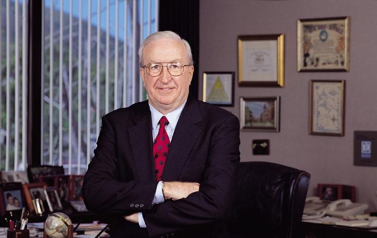 A smiling man with white hair and glasses, wearing a dark suit and red tie, stands with crossed arms in an office with framed pictures and a window.