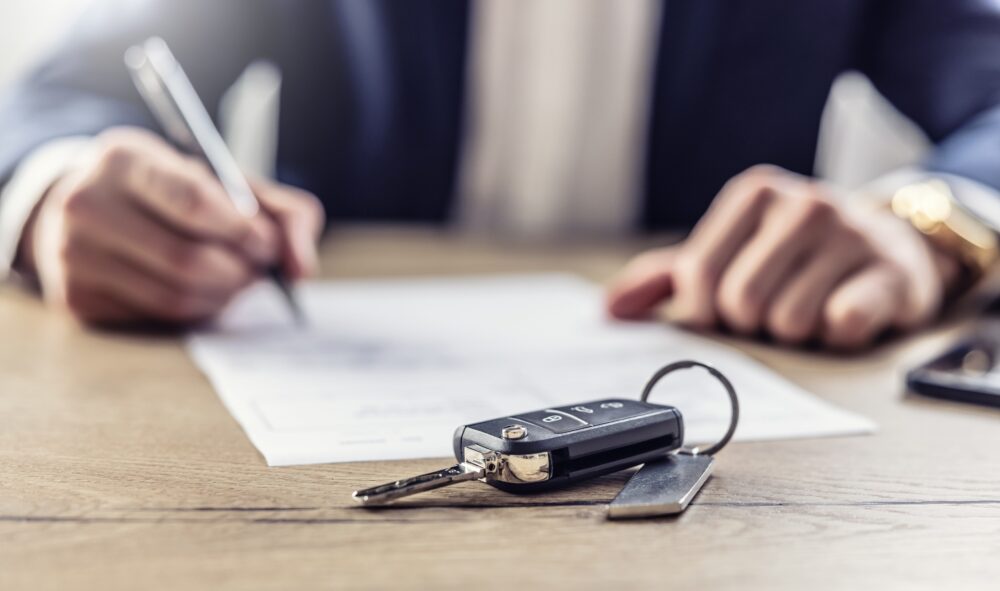 Agent signs a contract for the purchase or Leasing of a new car. New car keys in the foreground.