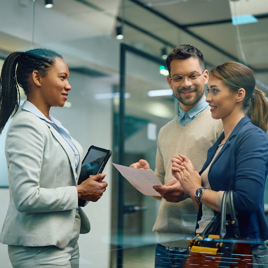 Three people, two women and one man, smiling and talking in an office. One woman holds a tablet, the man holds a document, and the other woman holds a handbag.
