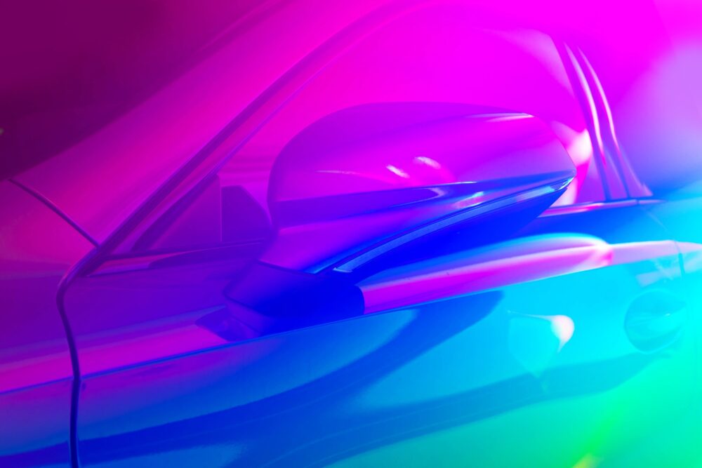 A close-up of a modern car side mirror and bodywork illuminated by vibrant blue and pink neon light.|