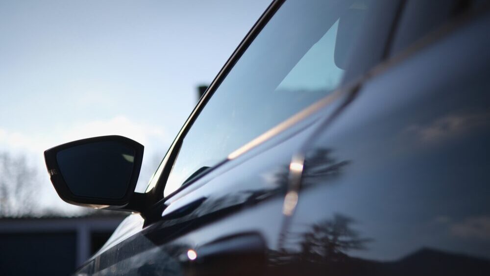 Close-up of a car’s side mirror and door with glossy reflections of the sky and trees, showcasing a sleek design, smooth curves, and tinted windows.