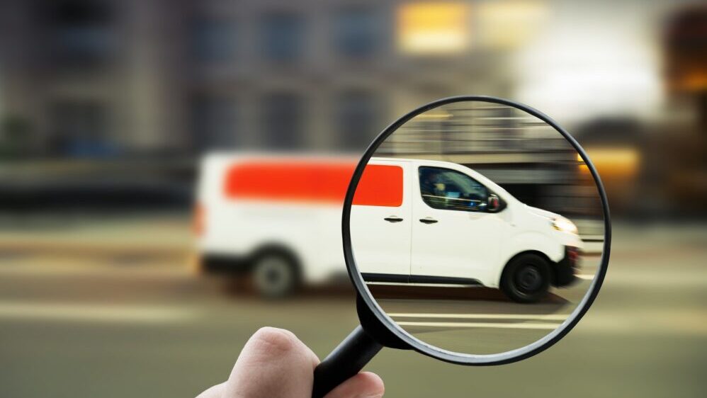 A magnifying glass focusing on a white delivery van driving through the city, representing logistics and urban transport.
